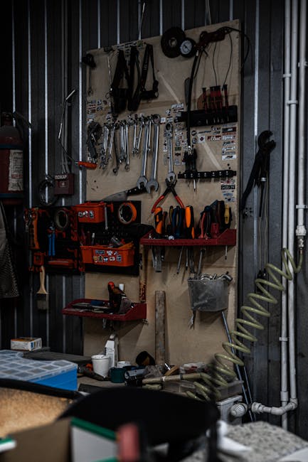 An interior view of a garage showing a concrete floor and exposed wooden ceiling beams with fluorescent lighting fixtures. Against the left wall, a white refrigerator is positioned next to a small, pa