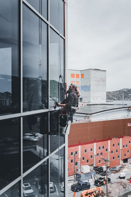 Two workers wearing helmets and safety harnesses are engaged in roof maintenance or repair atop a building structure. One worker is positioned on the ground, holding a rope or safety line secured to t