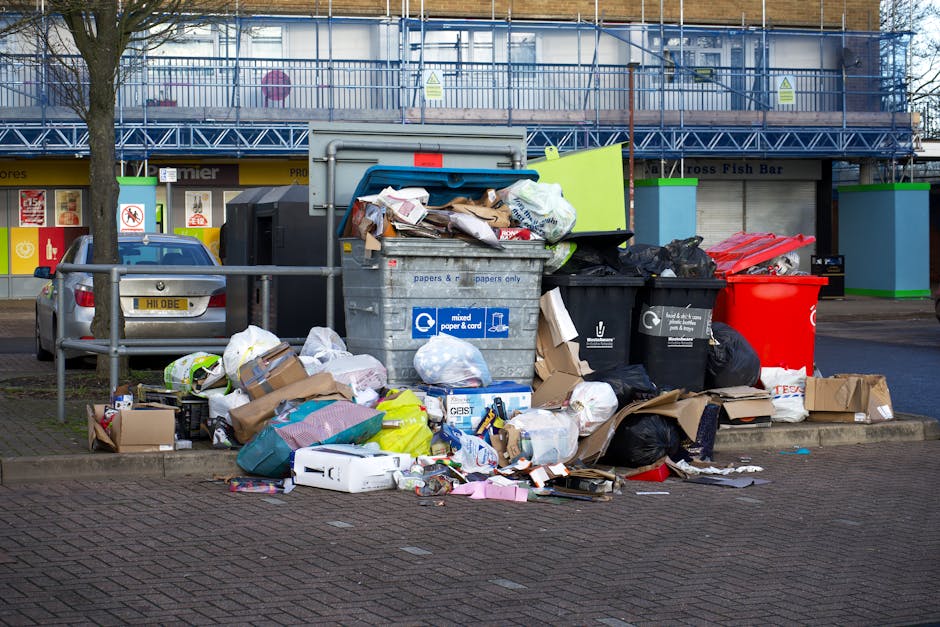 A large collection of mixed waste appears to be discarded outside a commercial property, piled loosely around multiple rubbish bins and bags. The waste includes flattened cardboard boxes, plastic pack