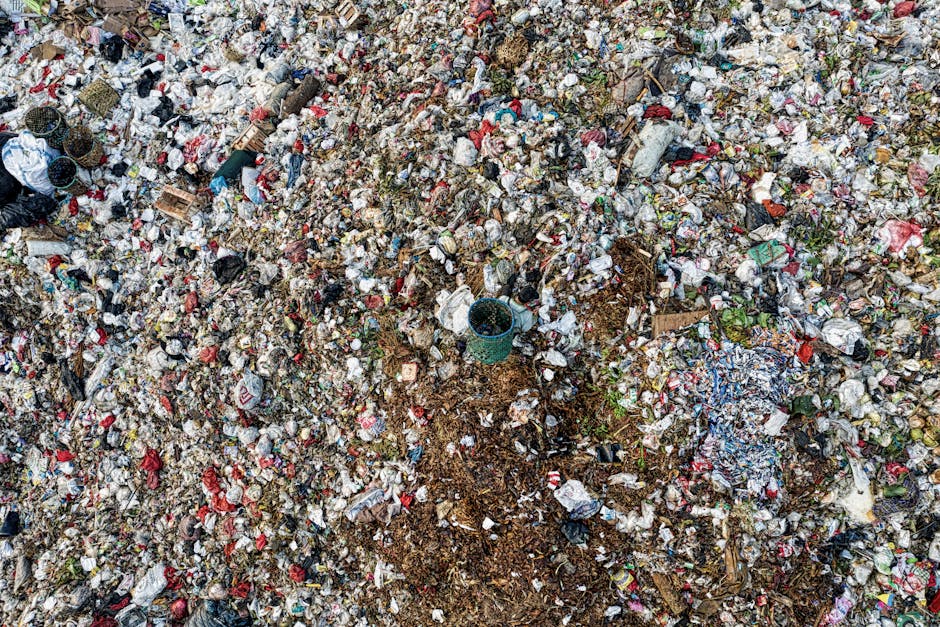 A small, cluttered area on the edge of a body of dark water, featuring a mixture of trash including plastic bottles, disposable cups, and paper debris scattered on muddy ground near a rocky embankment